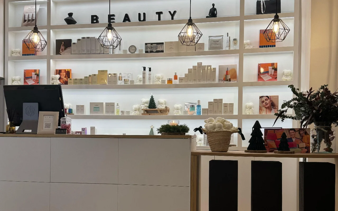 Un espace daccueil lumineux et moderne avec une grande étagère blanche rétroéclairée garnie de produits de soin. Des suspensions géométriques en fil noir pendent du plafond, et le comptoir daccueil est un meuble blanc épuré et minimaliste.