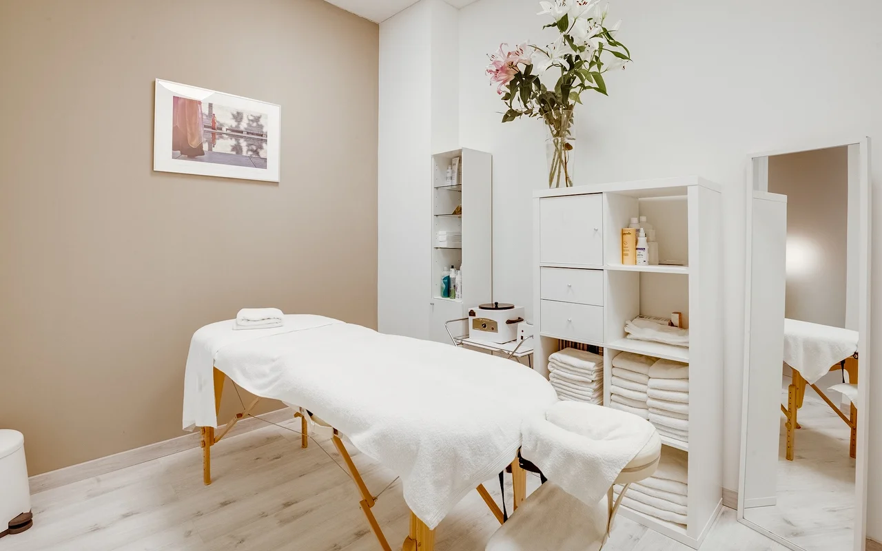 Une cabine de soin paisible avec une table de massage blanche recouverte de serviettes fraches. La pièce est dans des tons beige et blanc, avec un meuble de rangement blanc garni de serviettes pliées et un grand vase en verre avec des lys blancs et roses.