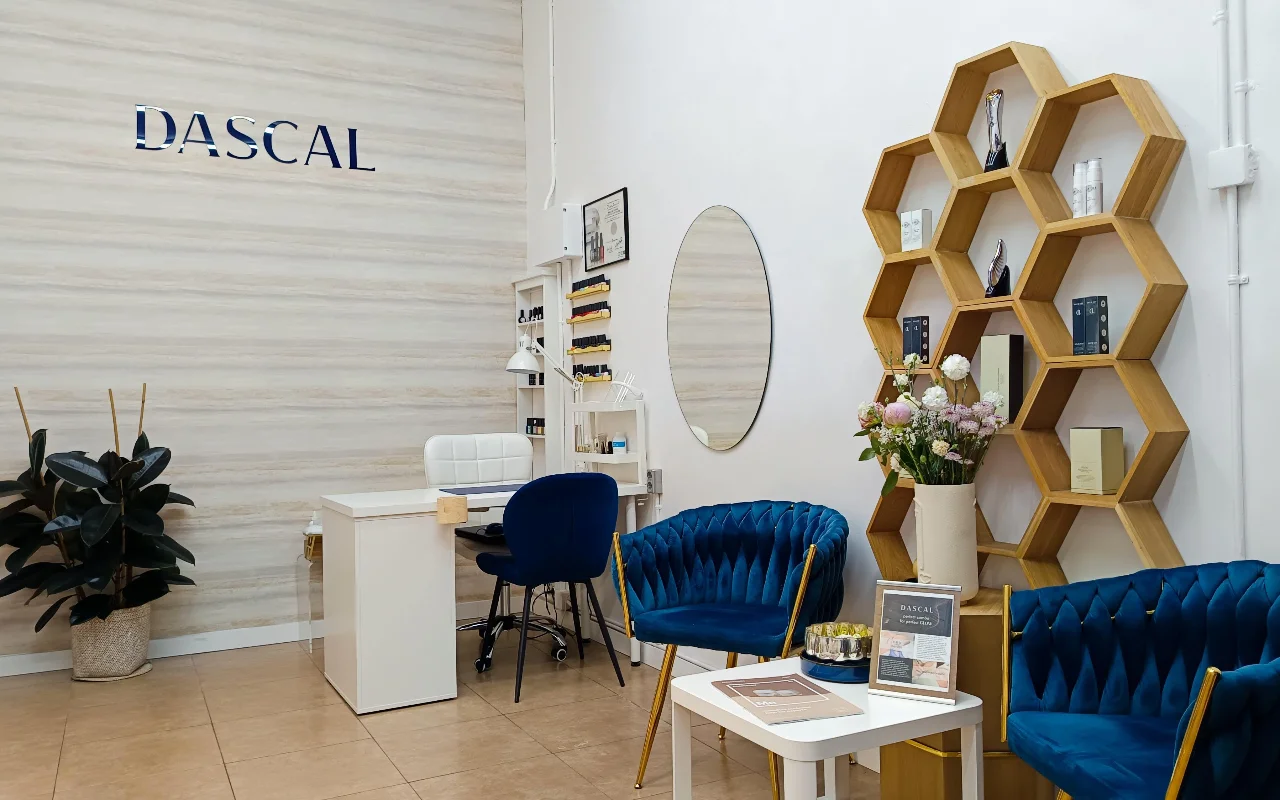 Un intérieur de salon de beauté élégant avec des fauteuils en velours bleu nuit et une étagère murale hexagonale en bois. Un bureau de manucure blanc est installé dans un coin contre un mur texturé clair, avec le mot DASCAL en lettres bleues.