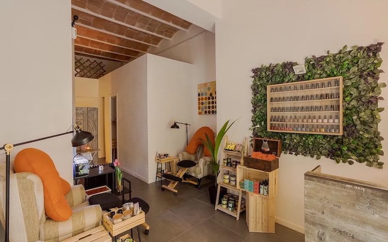 Un salon de manucure cosy au style bohème avec des fauteuils moelleux et des coussins orange. Le mur présente une grande installation végétale verticale entourant une vitrine en bois remplie de flacons de vernis à ongles.