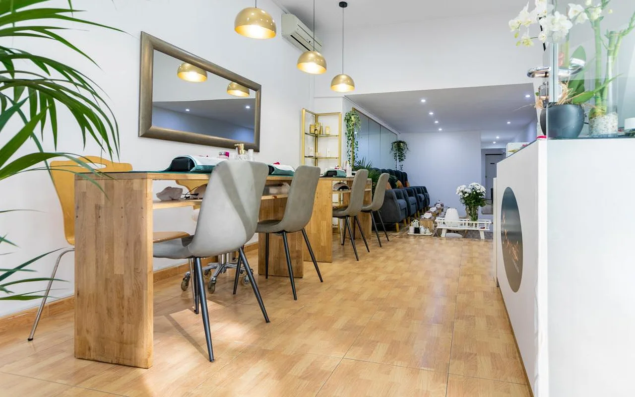 Un salon de manucure lumineux avec de longues tables en bois et des chaises grises modernes. Des suspensions dorées en dme sont accrochées au-dessus des postes, et un grand miroir à cadre foncé est fixé au mur blanc.