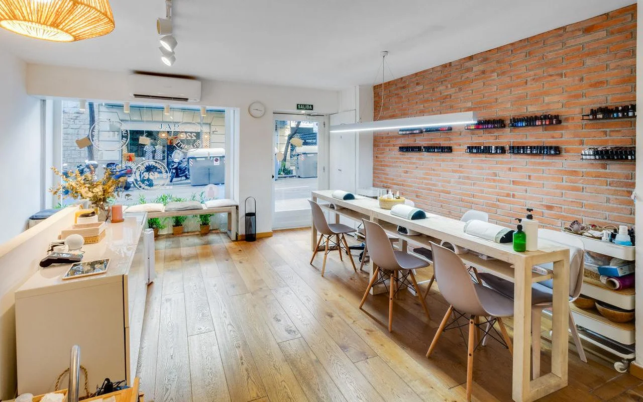 Un salon contemporain au style brut, avec un long comptoir de manucure blanc et un mur en briques rouges apparentes. De grandes fentres donnent sur une rue de la ville, inondant lespace au sol en bois de lumière naturelle.