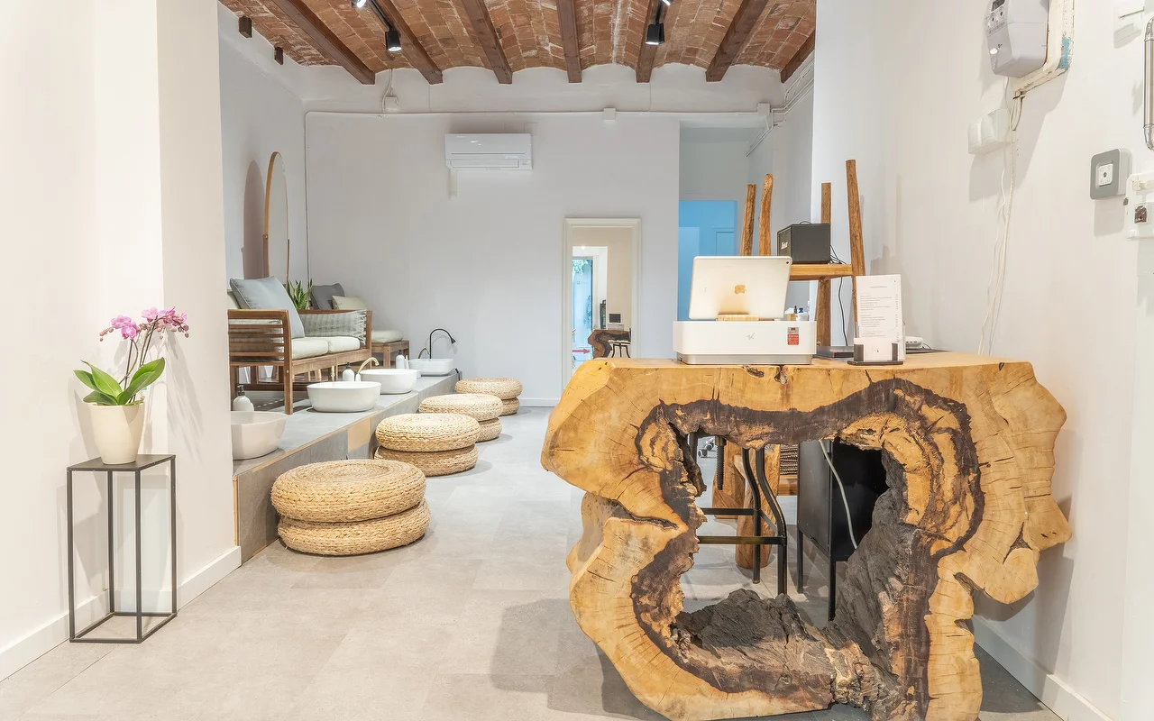 Un espace bien-tre minimaliste avec un comptoir daccueil en bois brut taillé dans un large tronc darbre. En arrière-plan, un poste de pédicure avec des coussins de sol tressés et des bassins blancs sous un plafond à poutres rustiques.