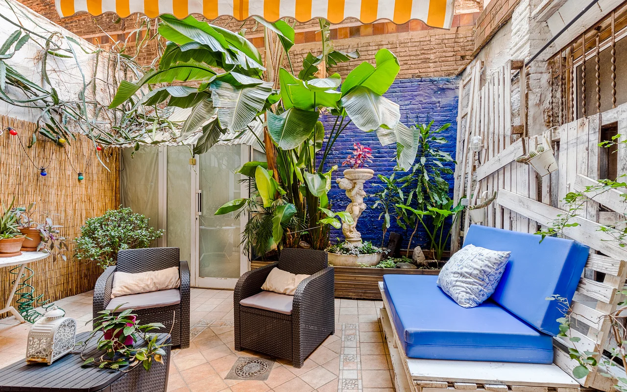 Un patio extérieur éclectique avec un mur daccent bleu vif et des plantes tropicales luxuriantes. On y trouve des fauteuils en osier foncé, une banquette à coussins bleus et un auvent à rayures jaunes et blanches.