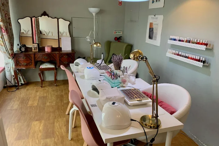 Vue intérieure d'un salon de beauté chaleureux spécialisé dans la manucure. Au premier plan, une longue table blanche est équipée de lampes UV, de lampes de bureau et de divers outils de prothésie ongulaire, entourée de chaises roses et blanches. En arrière-plan, une coiffeuse en bois vintage avec un miroir triptyque, un fauteuil vert et des étagères blanches présentant des rangées de flacons de vernis colorés.
