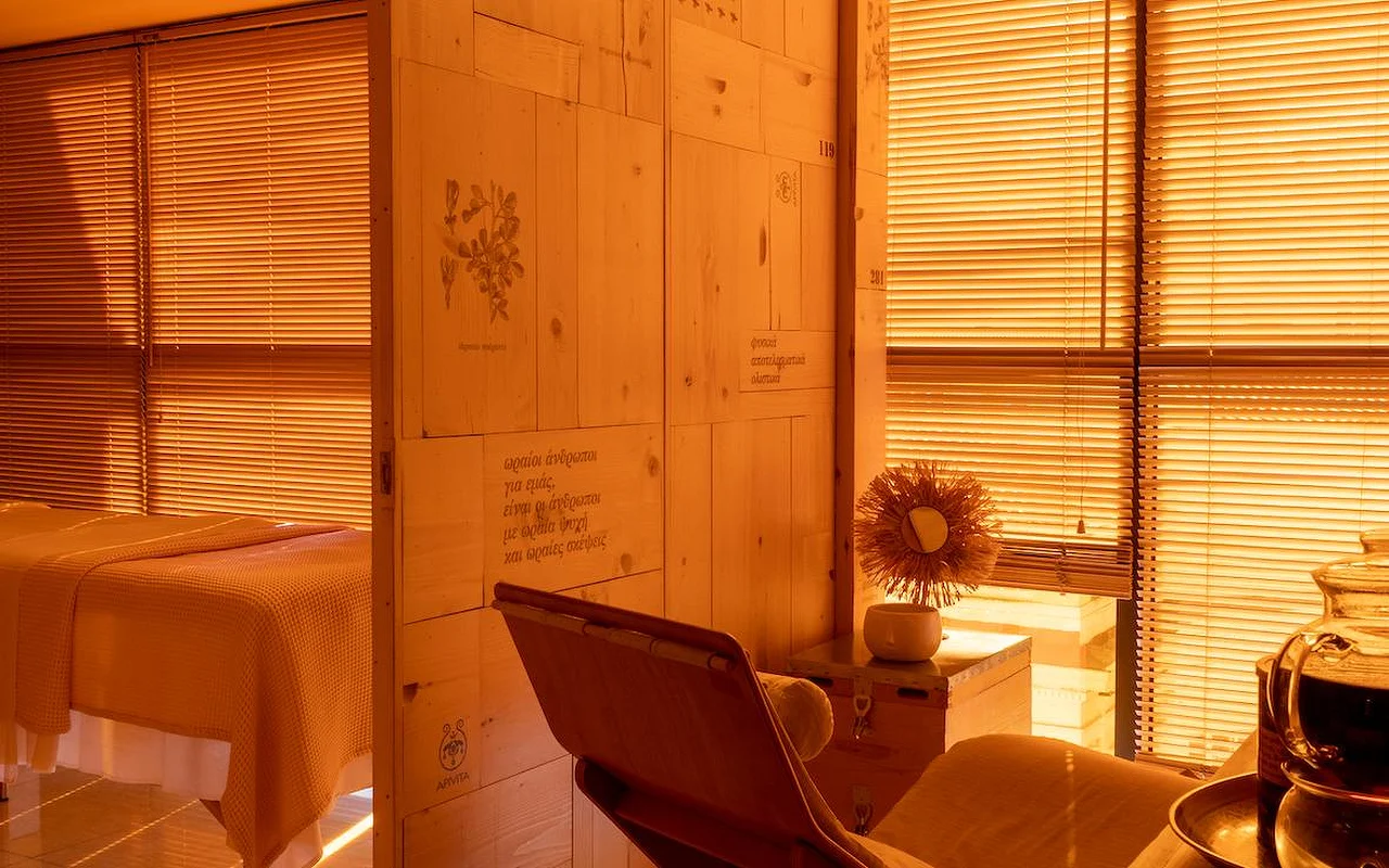 Une salle de soin spa chaleureuse baignée d’une lumière ambrée, avec des panneaux en bois naturel, des illustrations botaniques et du texte grec gravé sur le mur, un lit de soin drapé d’une couverture en nid d’abeille, et le logo APIVITA dans un coin.