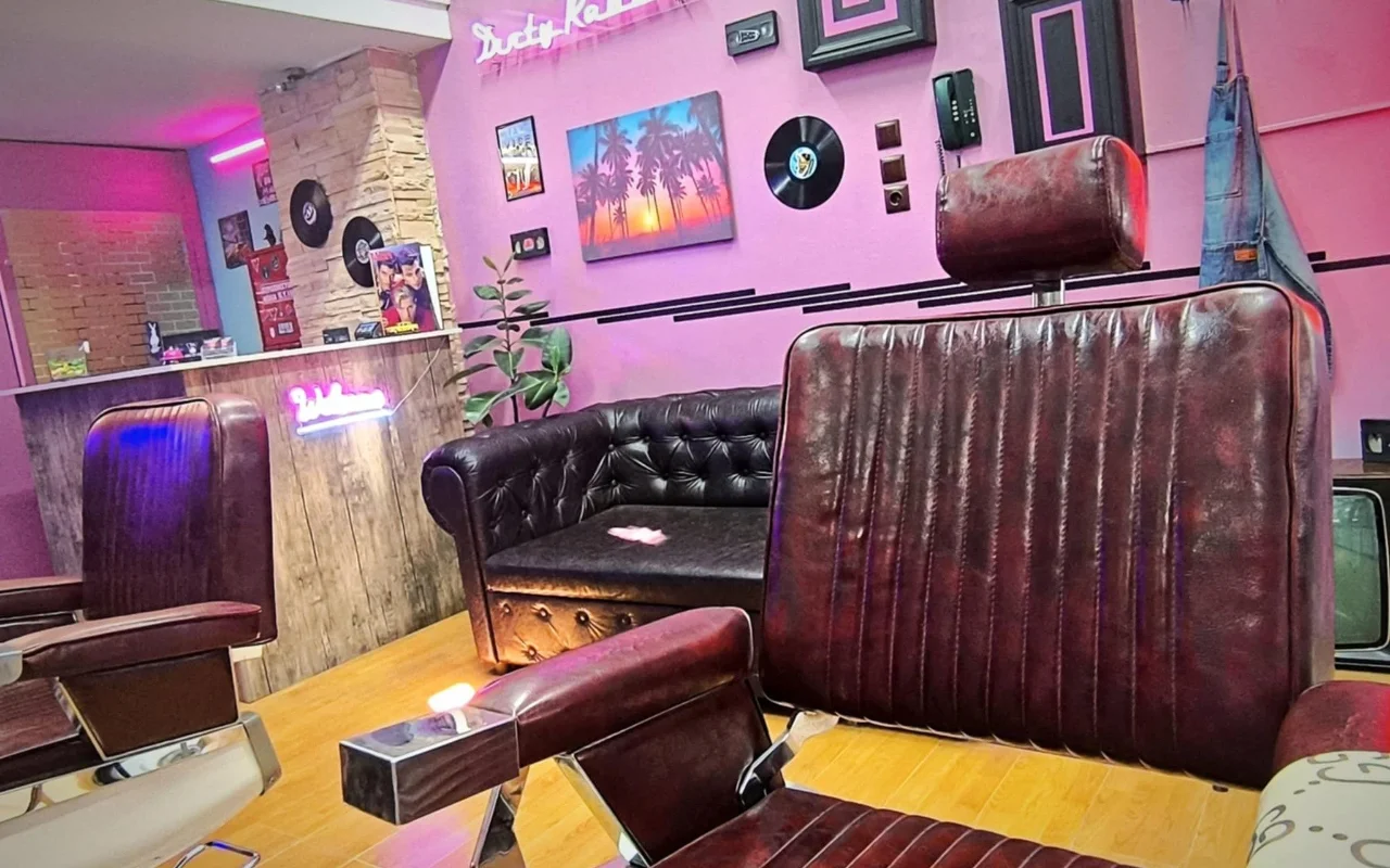 Un barbershop au style rétro avec des murs roses et violets, des enseignes néon, des vinyles accrochés en décoration, une peinture coucher de soleil tropical, des fauteuils de barbier en cuir foncé, et un canapé Chesterfield sur un parquet en bois chaleureux.
