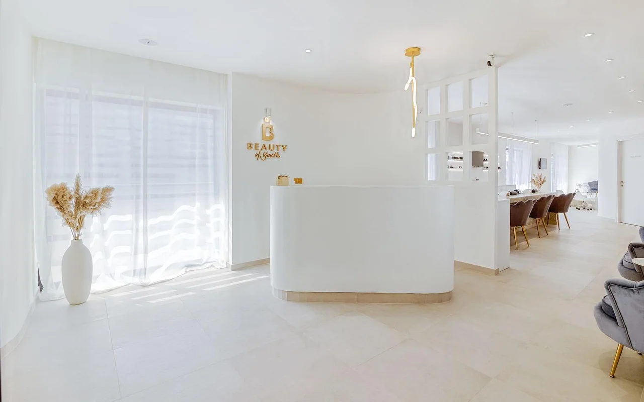 Un espace d’accueil lumineux, entièrement blanc, avec un comptoir arrondi, le logo doré Beauty of Youth au mur, une suspension sculpturale, des rideaux voilés filtrant la lumière naturelle, et des fauteuils de soin en velours brun visibles en arrière-plan.
