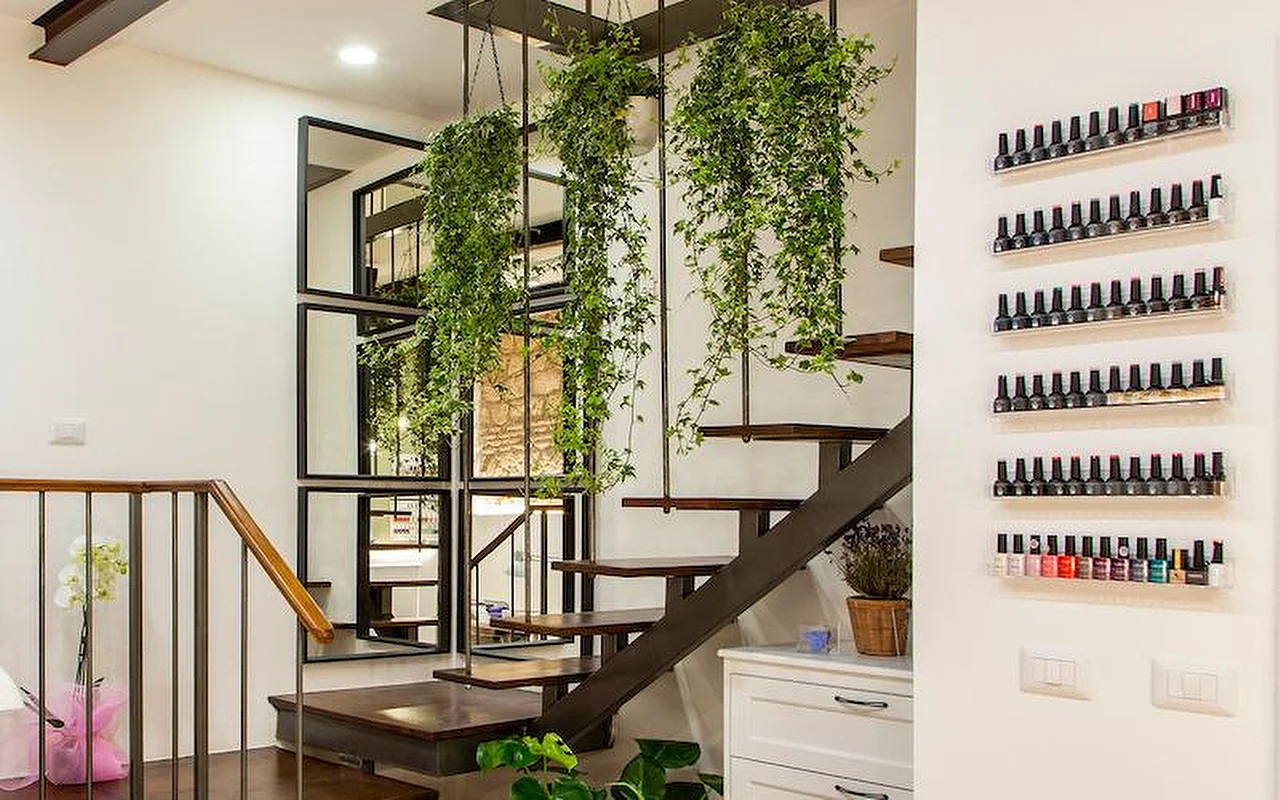 Un intérieur moderne et élégant avec un escalier en porte-à-faux en bois et métal. Des plantes retombantes luxuriantes pendent du plafond entre les marches, et des rangées de flacons de vernis colorés sont exposés sur des étagères transparentes fixées au mur blanc.