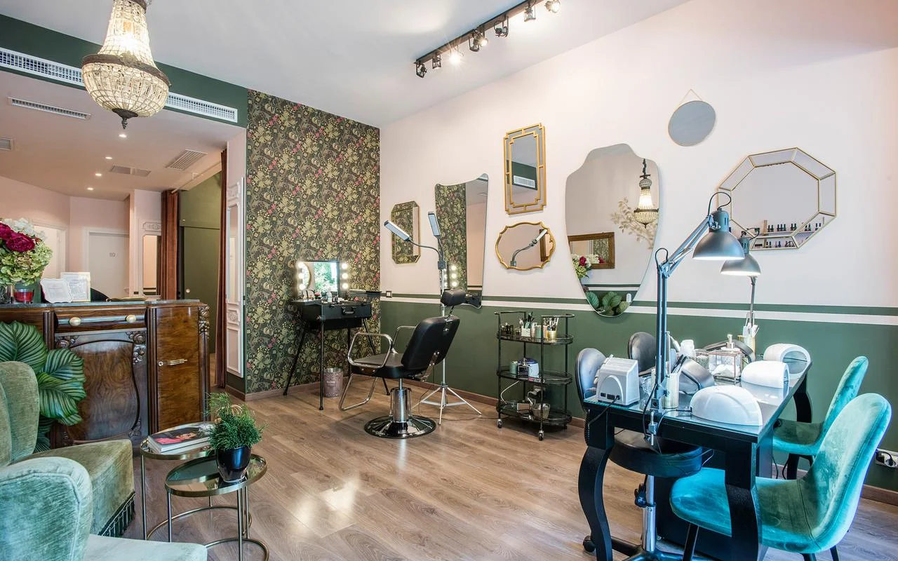 Un salon au charme vintage avec des murs blancs et vert forêt. Il présente un papier peint à motifs floraux, un assortiment de miroirs dorés aux formes irrégulières, un poste de maquillage professionnel et des assises en velours vert.