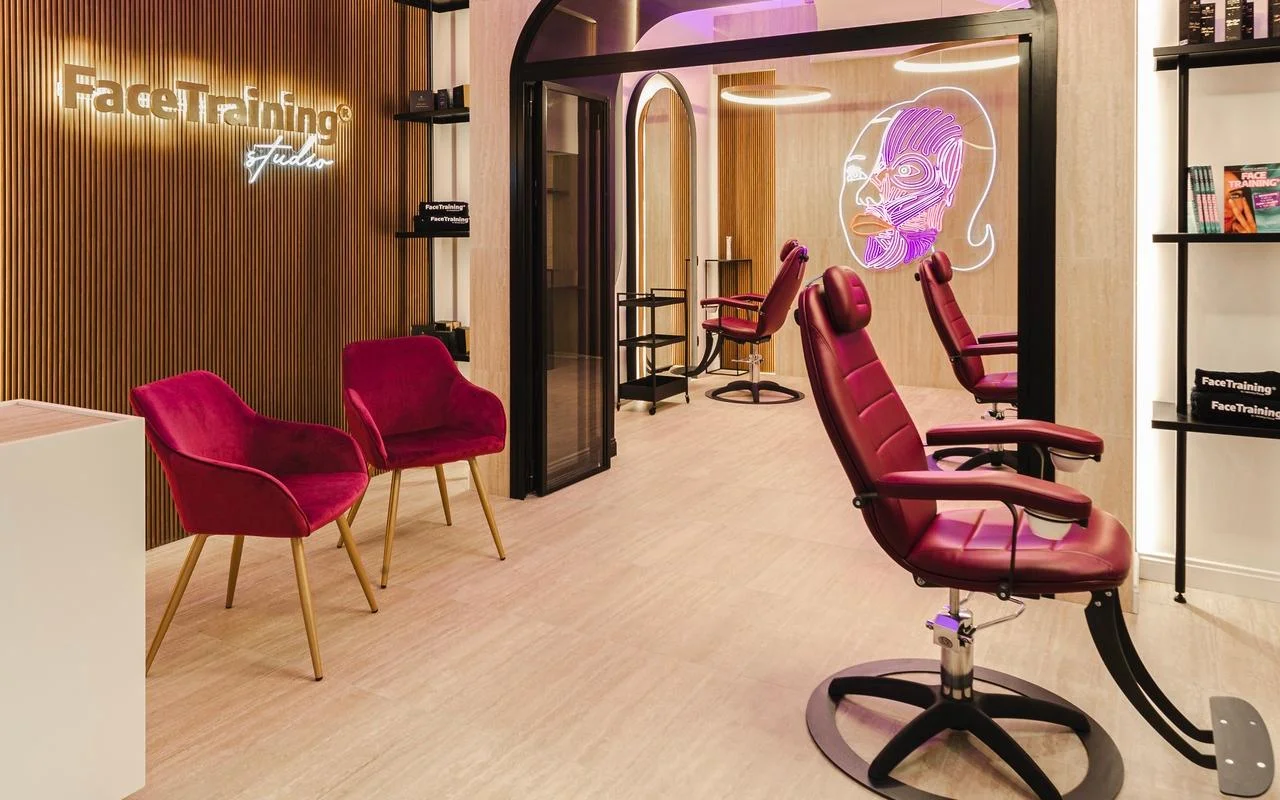 Un studio minimaliste avec un mur à lattes de bois et un logo rétroéclairé. L’espace de soin comprend des fauteuils professionnels en cuir bordeaux et une installation néon représentant un diagramme stylisé des muscles du visage sur une cloison vitrée.