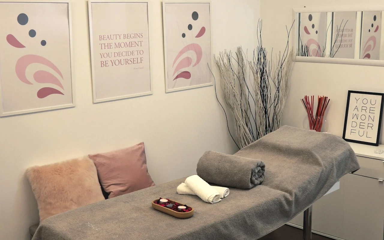 Une cabine de soin lumineuse avec une table de massage recouverte d’une serviette grise. Le mur est décoré d’art abstrait aux tons pastel et de citations inspirantes, tandis que des branches blanches décoratives et des bougies créent une atmosphère apaisante.