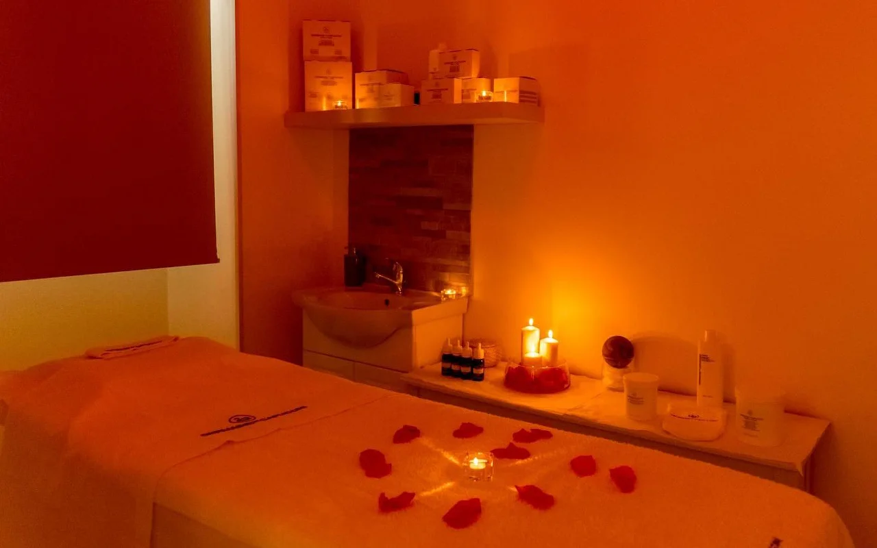 Une cabine de massage baignée d’une lumière orangée chaleureuse et de bougies vacillantes. Des pétales de rose rouges sont disposés en cercle autour d’une bougie centrale sur la table de soin, avec des produits de beauté soigneusement rangés sur une étagère à l’arrière-plan.