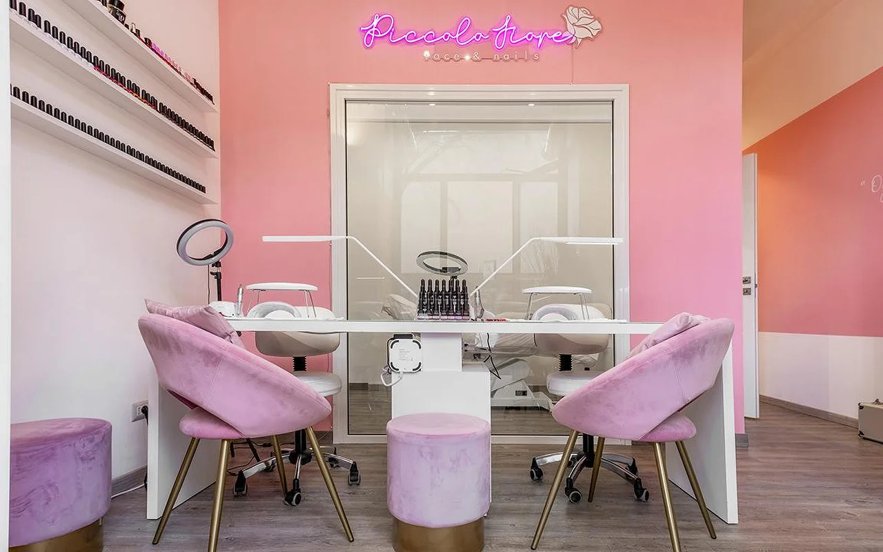 Un poste de manucure aux murs rose vif, avec une enseigne néon rose fuchsia. Des fauteuils en velours lilas aux pieds dorés entourent un bureau blanc minimaliste équipé de lampes LED professionnelles pour les ongles.