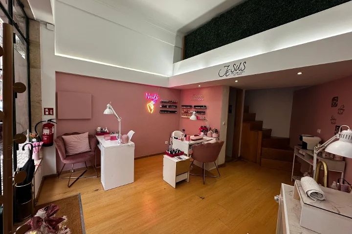 Un studio d'ongles chic aux murs rose pâle et au parquet chaleureux. La pièce contient plusieurs bureaux de manucure blancs associés à des fauteuils en velours rose poudré. Une enseigne néon rose vif en forme de main tenant un flacon de vernis avec le mot « Nails » brille sur le mur, et des rangées de vernis sont exposées sur de petites étagères. Une mezzanine avec un mur végétal est visible au-dessus.