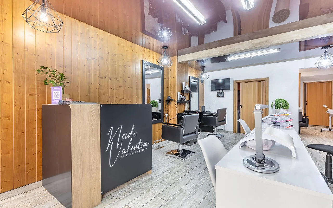 Un salon de beauté contemporain avec un comptoir d'accueil à gauche portant la marque « Neide Valentim Instituto de Beleza ». Les murs sont habillés de panneaux verticaux en bois chaleureux et de grands miroirs. Des fauteuils de coiffure noirs se dressent devant les miroirs, tandis qu'un poste de manucure blanc avec des chaises blanches longe le côté droit du sol en carrelage gris.