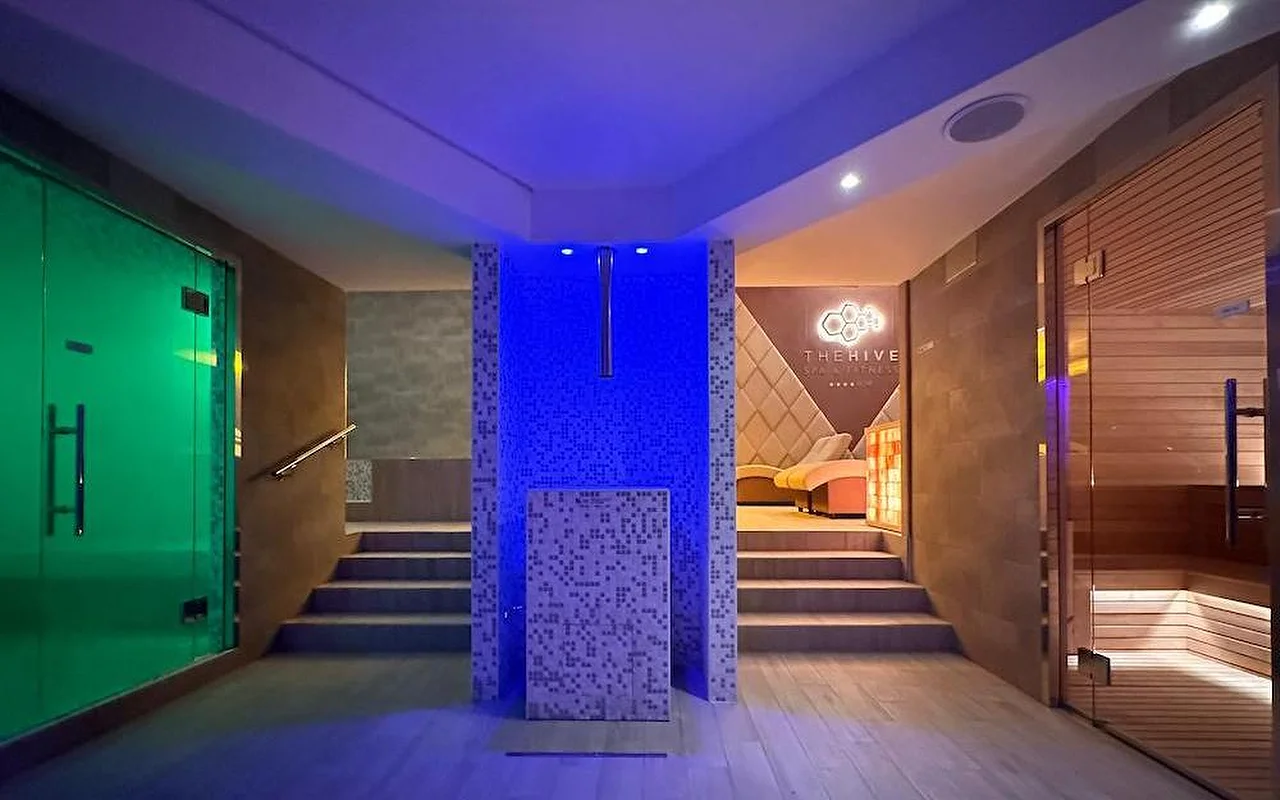 L’espace thermal d’un spa de luxe avec un éclairage atmosphérique bleu et vert. L’espace comprend un sauna finlandais en bois, l’entrée d’un hammam et un élément central en mosaïque avec des marches menant aux zones de détente.