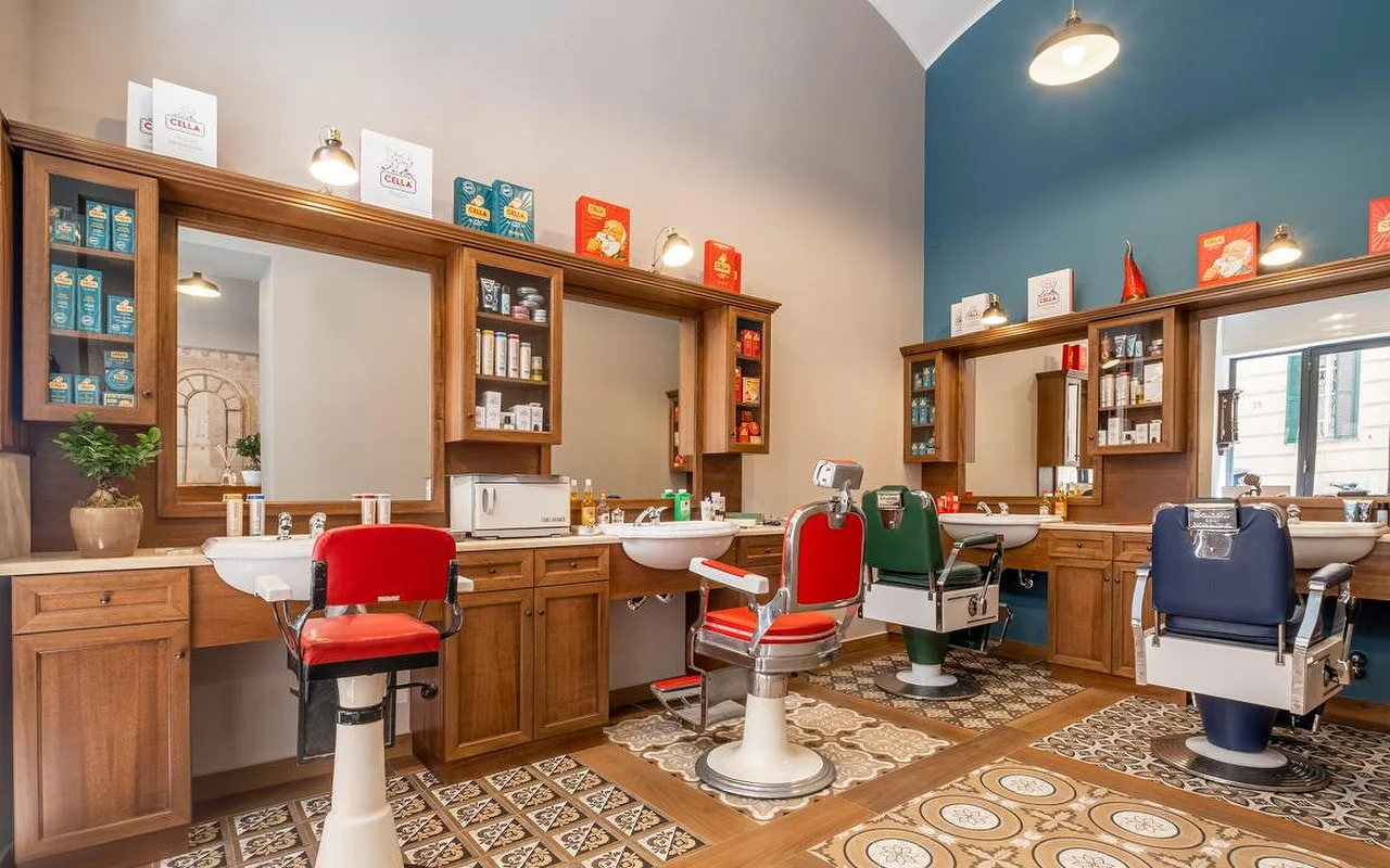 Un barbier haut de gamme de style traditionnel, avec des meubles en bois riche, un sol à carreaux vintage et des fauteuils de barbier professionnels en rouge, vert et bleu marine. Des produits de rasage traditionnels sont exposés sur les étagères.