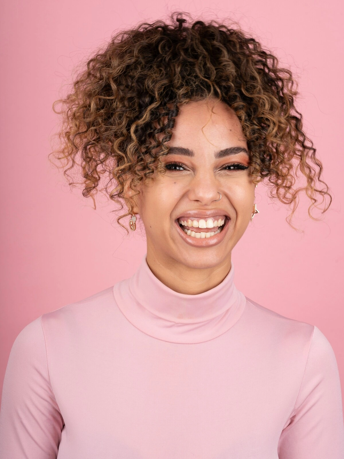 Photo studio d'une femme souriante avec un balayage sur cheveux afro, vêtue d'un col roulé rose pâle