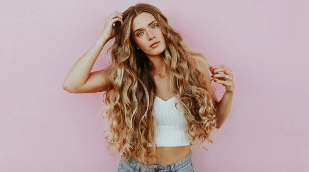 Photo studio d'une femme aux très longs cheveux ondulés effet soleil réalisés en balayage, vêtue d'un jean bleu clair et d'un top court en maille blanc
