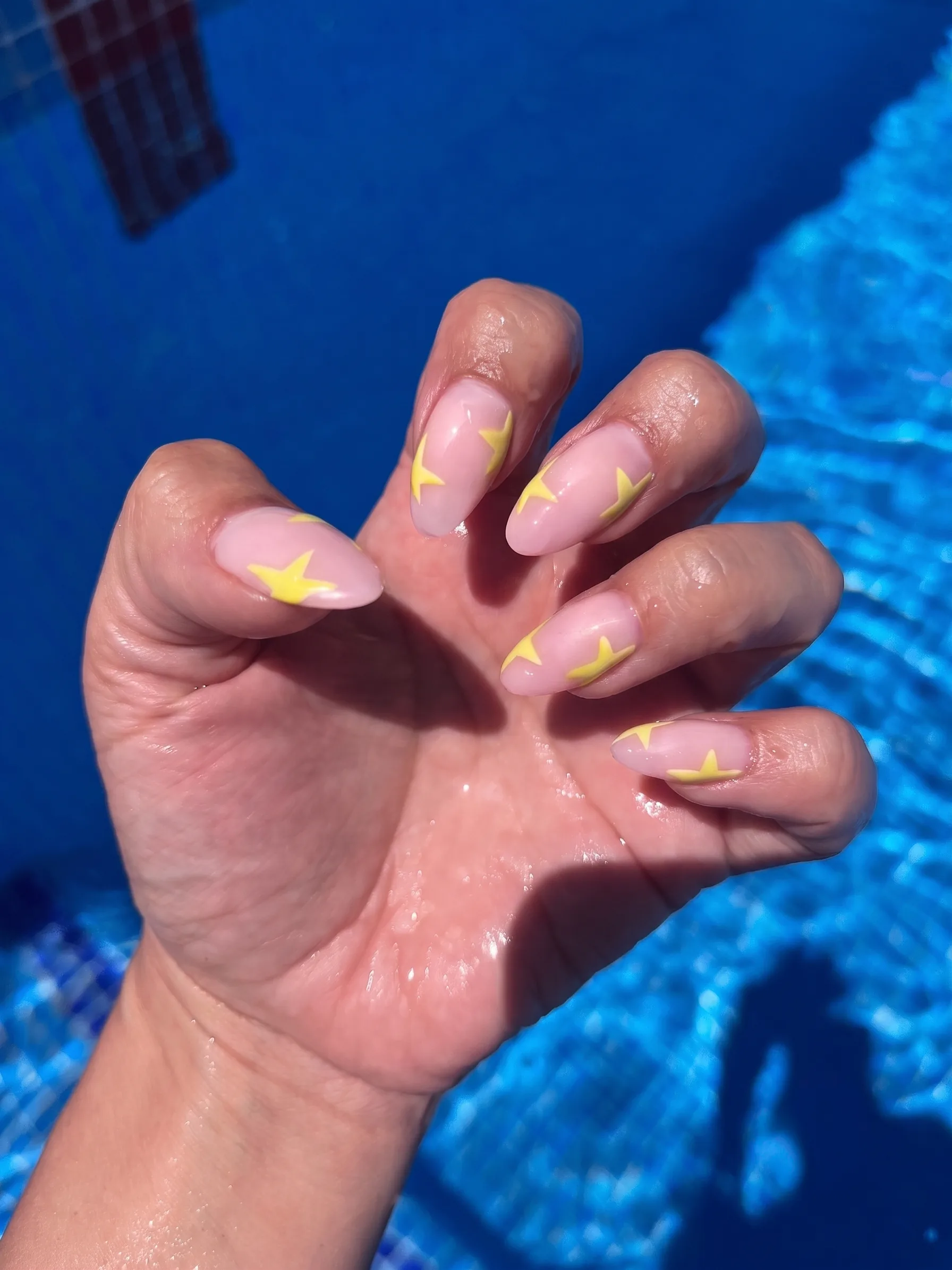 Ongles en amande nude transparent rehaussés d'étoiles jaune beurre, photographiés au bord de la piscine.