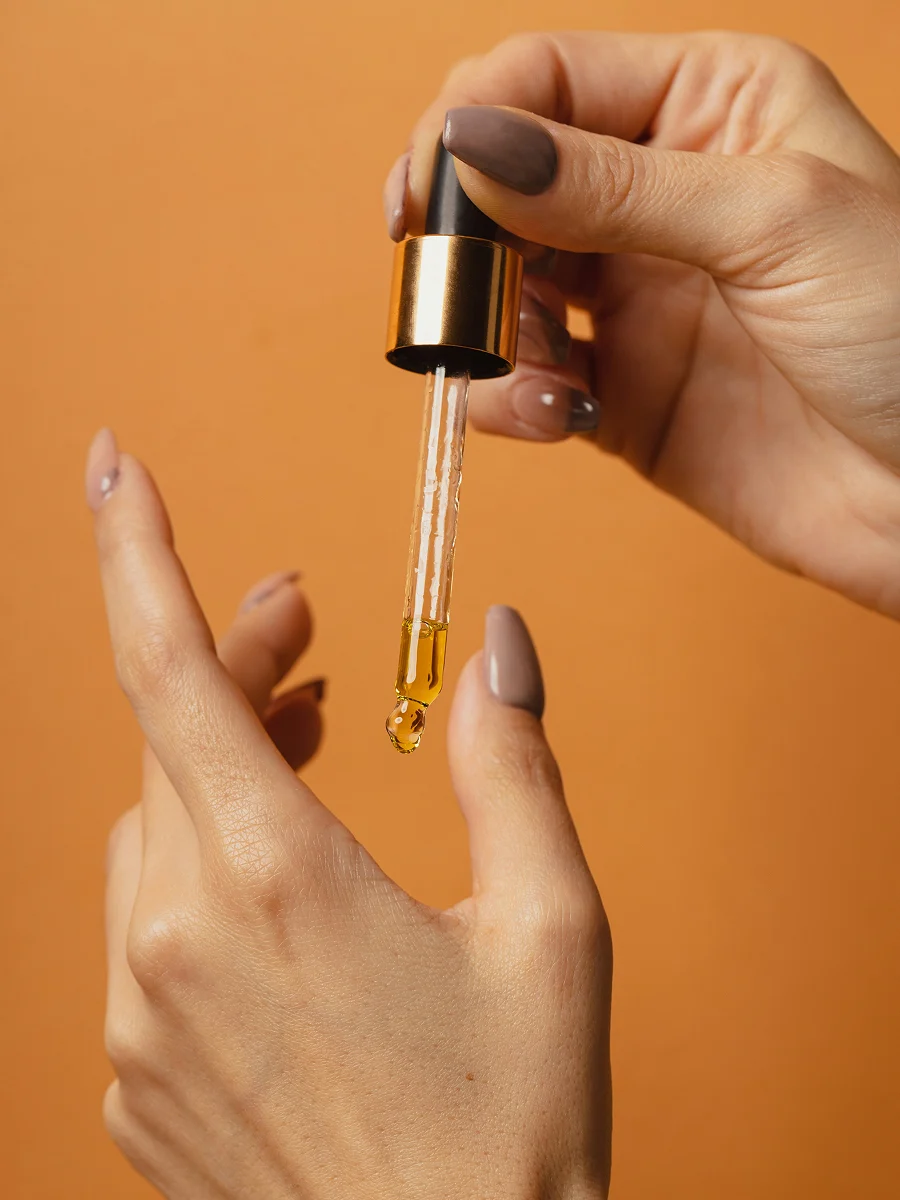 Gros plan de deux mains manucurées, l’une tenant une pipette en verre d’où s’écoule de l’huile pour cuticules