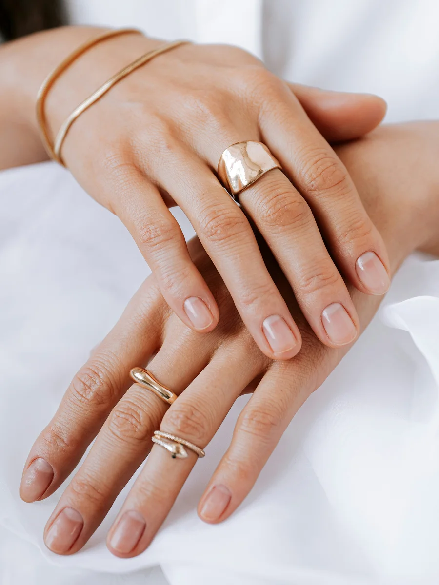 Gros plan de deux mains posées sur un drap blanc, aux ongles naturels et sains, ornées de bagues et de bracelets en or rose