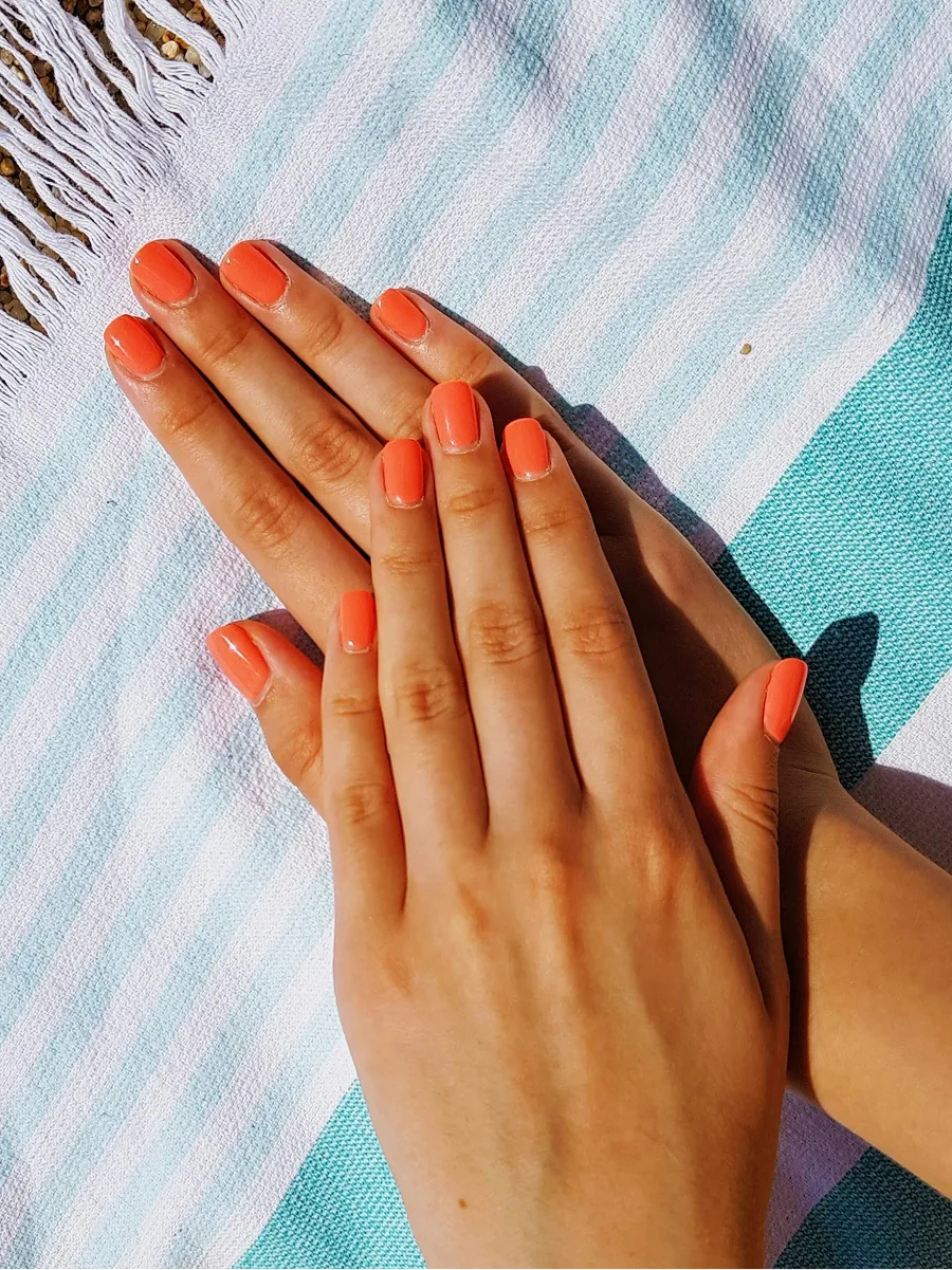 Vue du dessus de deux mains à la manucure gel orange brillante, posées sur une serviette de plage à rayures bleues et blanches