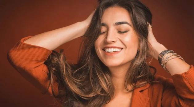 Gros plan d'une femme souriante, les yeux fermés, passant les mains dans ses longs cheveux châtains et doux