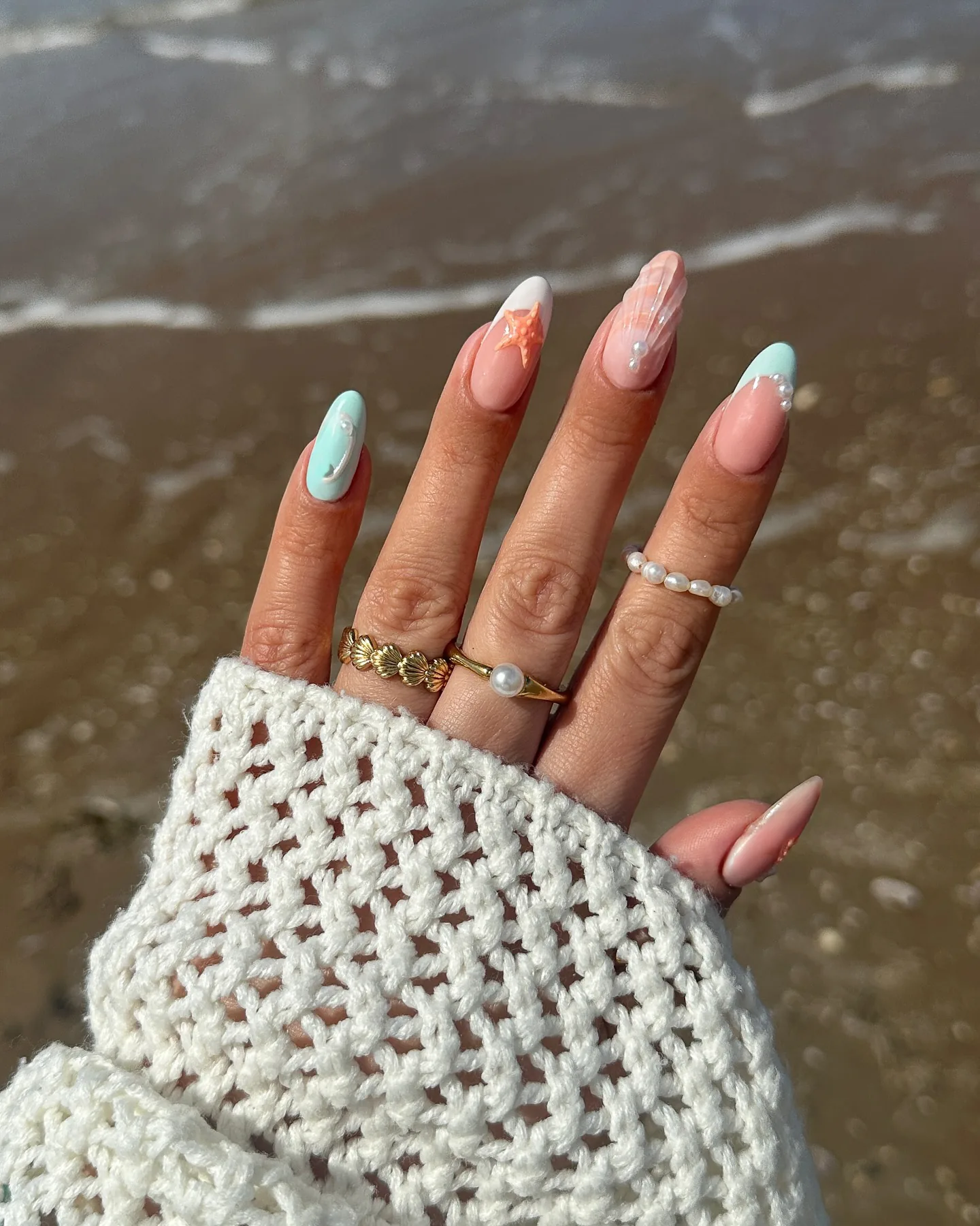 Ongles en amande ornés d'un nail art sirène pastel – une étoile de mer, des accents nacrés et un éclat irisé – sur du sable humide.