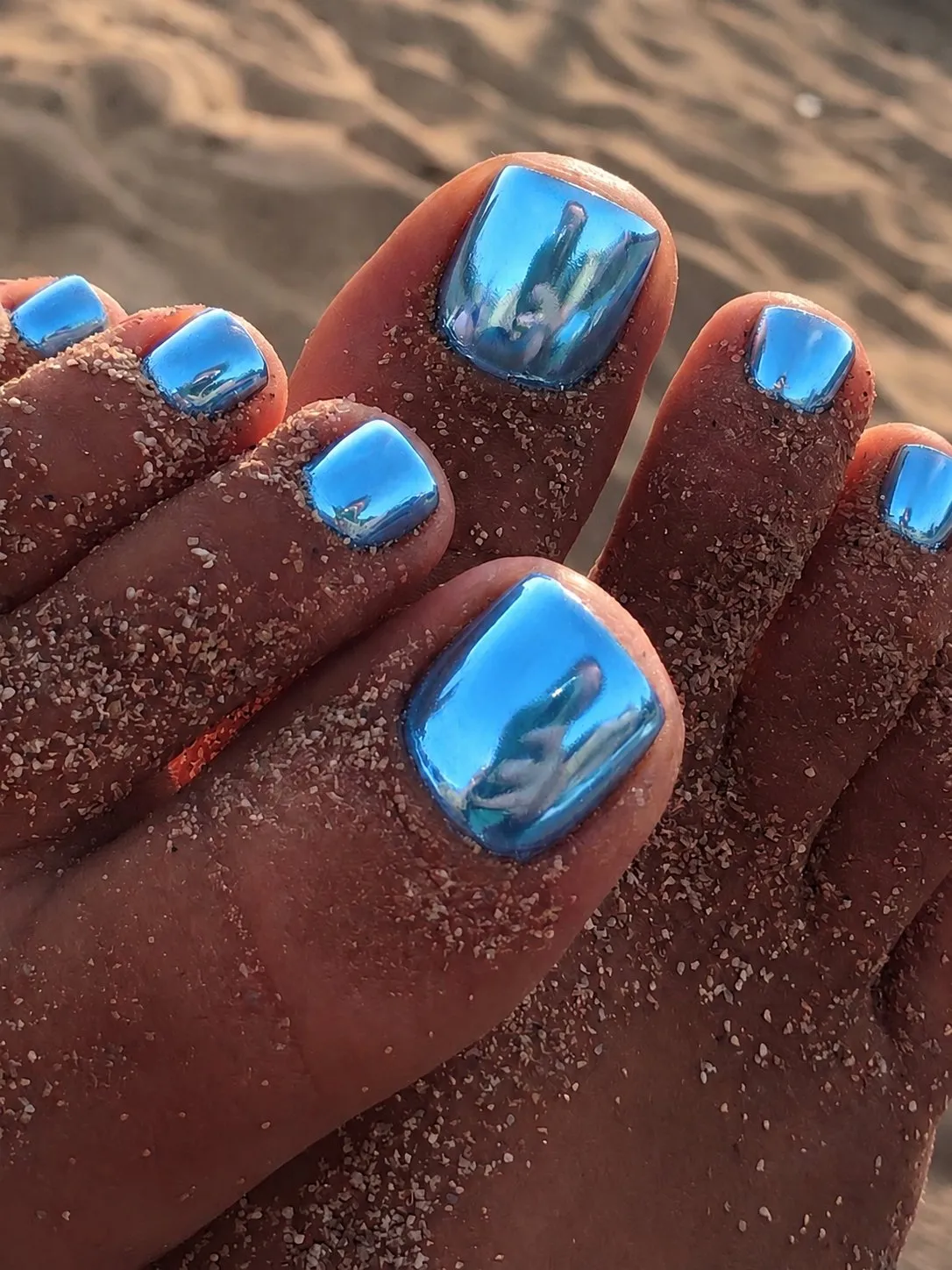 Pédicure chromée bleu océan métallisée sur des pieds dans le sable, à la plage