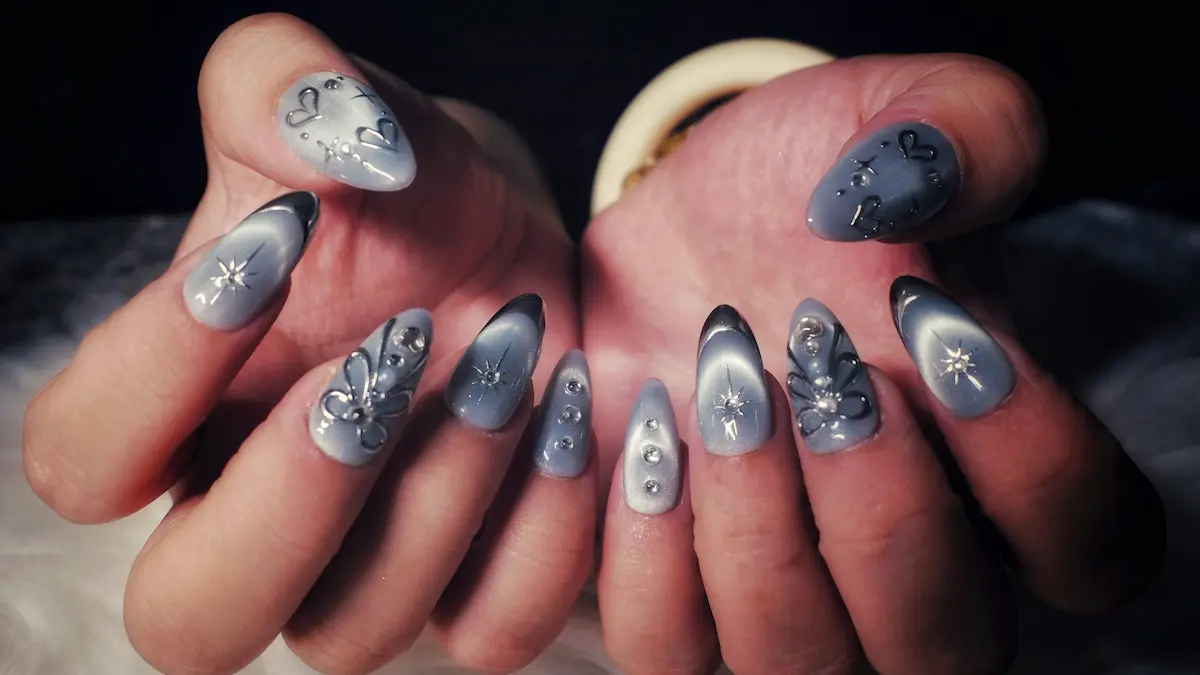 Gros plan sur les mains d'une femme arborant des ongles en résine sophistiqués, motif œil-de-chat gris, cœurs et étoiles argentés, et décorations en pierres scintillantes