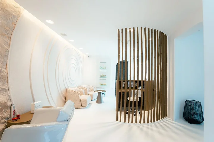 Un intérieur de spa calme aux allures de grotte, aux murs blancs incurvés, dont l'un est sculpté en ondulations concentriques, avec un éclairage indirect doux, des fauteuils matelassés crème et un paravent en lattes de bois courbé délimitant un coin soin tranquille.