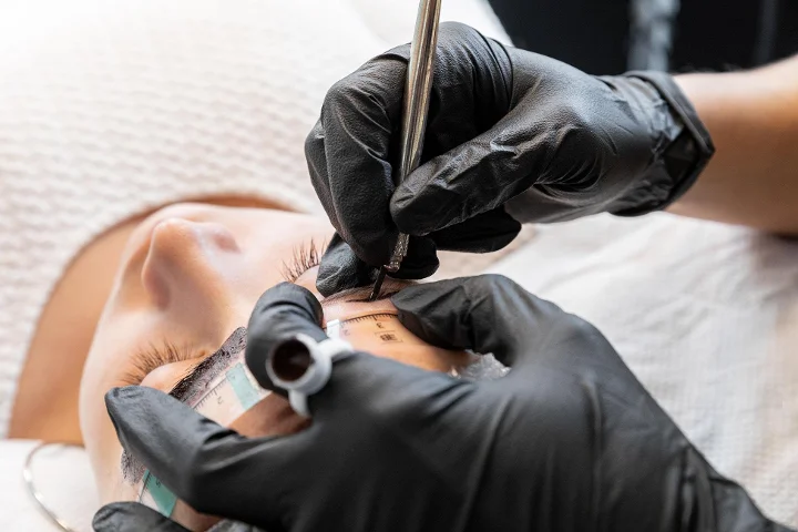 Gros plan sur un soin des sourcils en cours : une praticienne en gants noirs trace et marque le sourcil d'une cliente allongée à l'aide d'un compas et d'un crayon, les yeux fermés.