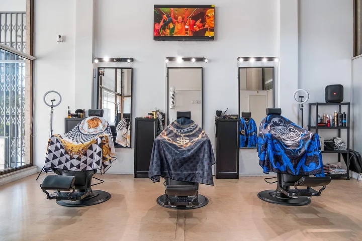 Un salon de barbier lumineux sous haut plafond, avec trois fauteuils noirs habillés de capes à motifs, chacun face à un miroir cerné d'ampoules, des anneaux lumineux, un écran mural et de hautes fenêtres.
