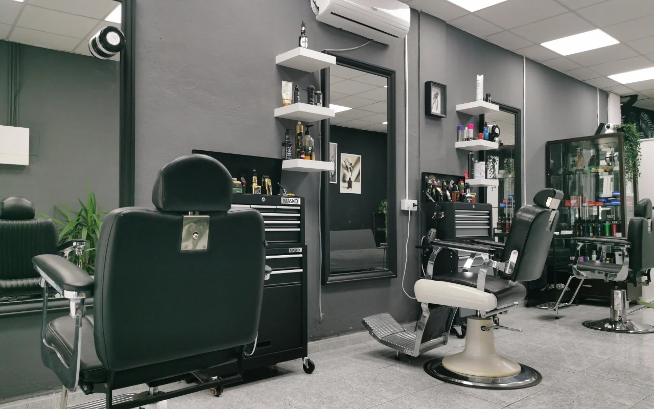 Un barbier traditionnel aux murs gris anthracite, des fauteuils de barbier classiques en cuir noir à bases et repose-pieds chromés, des étagères flottantes blanches garnies de produits de soin, et de grands miroirs sur un sol carrelé.