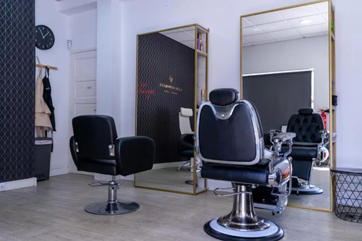 Un intérieur de salon épuré avec un fauteuil de coiffure noir et un fauteuil de barbier en chrome et cuir sur un parquet clair, un grand miroir à cadre doré, une horloge murale et des blouses suspendues près d'un mur d'accent sombre.