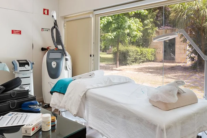Une cabine de soins avec une table de massage drapée de blanc près d'une baie vitrée donnant sur un jardin verdoyant, un appareil de soins corporels sur pied, une lampe-loupe et une table d'appoint en verre garnie de produits.