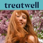 Portrait en gros plan d'une jeune femme rousse souriante avec une frange, dont les cheveux bougent au vent devant un arrière-plan de fleurs de lilas roses. Un bandeau bleu clair en haut affiche le logo "Treatwell" en bleu foncé.