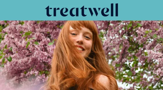 Portrait en gros plan d'une jeune femme rousse souriante avec une frange, dont les cheveux bougent au vent devant un arrière-plan de fleurs de lilas roses. Un bandeau bleu clair en haut affiche le logo "Treatwell" en bleu foncé.