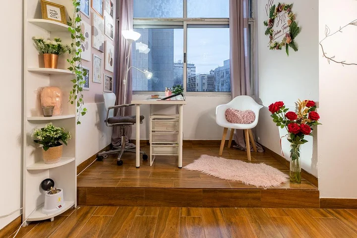Un petit studio d'esthétique sur une estrade en bois, avec un bureau blanc et une lampe à pince, une chaise de bureau et un fauteuil, des étagères d'angle garnies de plantes, un vase de roses rouges et une fenêtre donnant sur la ville.