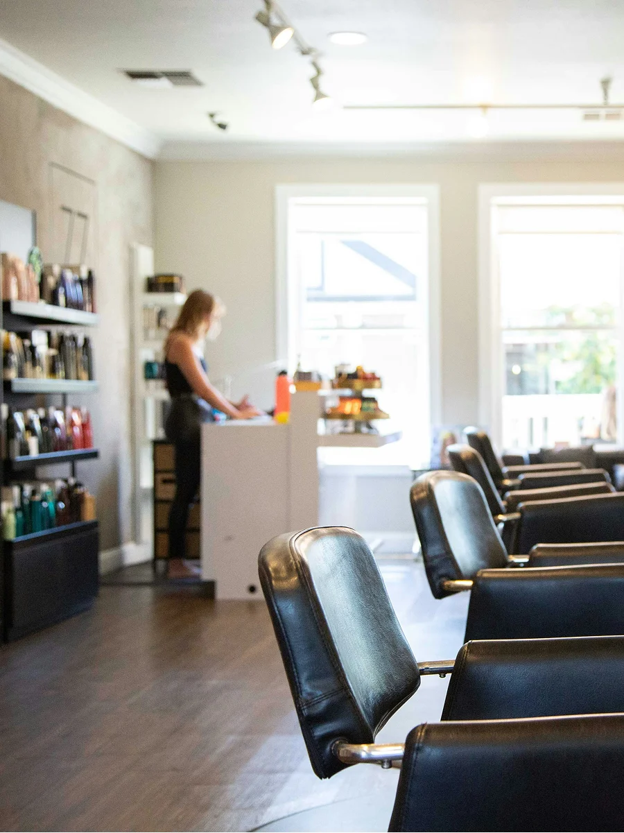 Vue intérieure d’un salon de coiffure propre, lumineux et bien aéré, avec de grandes fenêtres, des fauteuils noirs et des étagères de produits