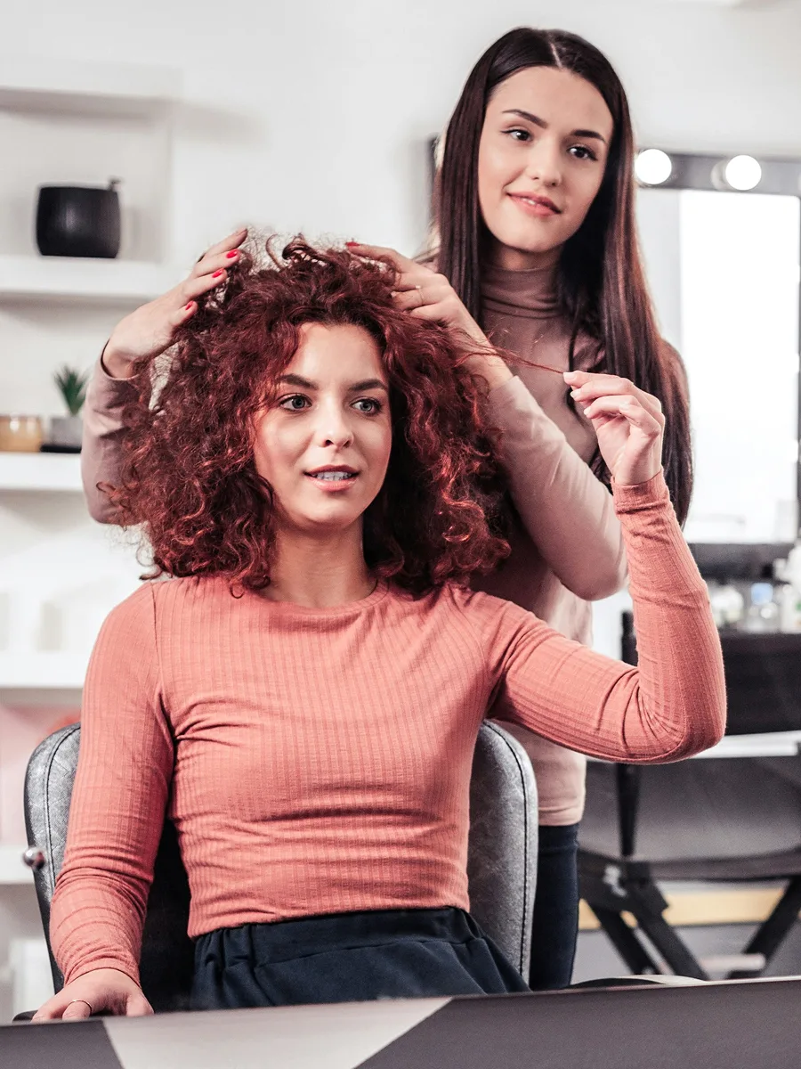 Une cliente aux cheveux bouclés teints en roux est assise chez le coiffeur pendant que son styliste discute de son soin