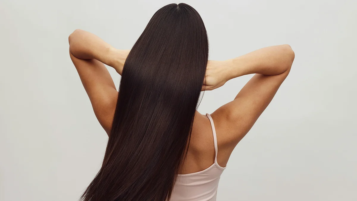 Vue de dos d’une femme en haut blanc à fines bretelles, les bras levés, passant les doigts dans ses longs cheveux bruns brillants
