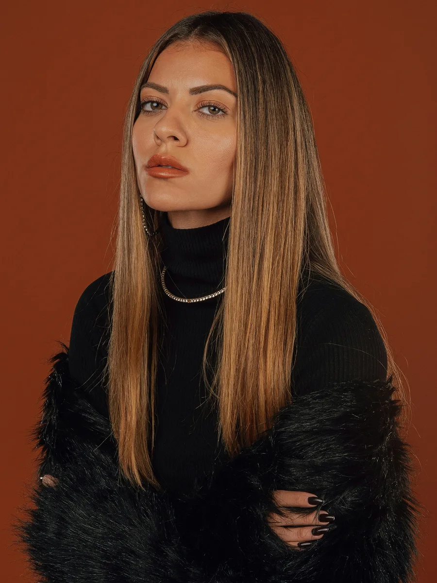 Portrait en studio d’une femme aux longs cheveux châtains brillants, vêtue d’un haut noir, d’une étole en fourrure noire et de créoles et d’un collier scintillants