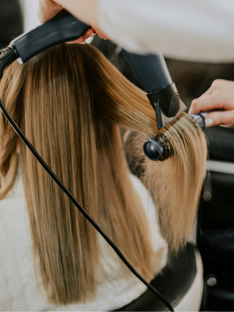 Vue de dos d’une femme chez le coiffeur pendant que le styliste lui sèche les cheveux bien lisses lors d’un rendez-vous de lissage à la kératine