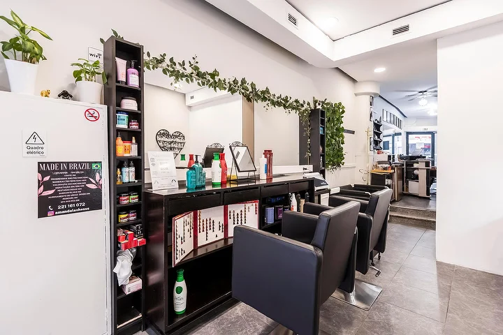 Un salon de coiffure tout en longueur avec des fauteuils noirs alignés le long d'un mur de miroirs, une étagère sombre garnie de produits capillaires, une guirlande de verdure retombant sur le miroir et une enseigne Made In Brazil encadrée sur une colonne blanche.