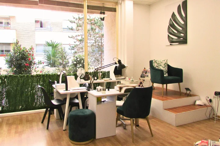 Un espace manucure lumineux avec deux tables à ongles blanches et leurs lampes, des fauteuils en velours vert et un tabouret assorti, une toile à feuille de monstera au mur et de grandes fenêtres donnant sur une terrasse plantée.