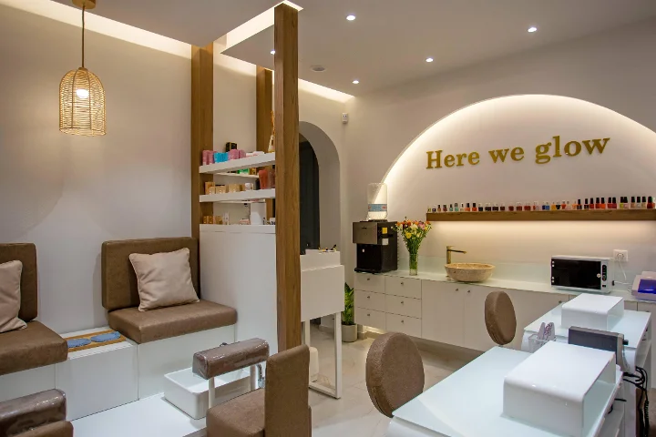 Un salon d'ongles chaleureux à la lumière douce, avec une suspension en rotin tressé, des banquettes de pédicure brunes rembourrées, des tables de manucure blanches, une étagère de vernis colorés et une enseigne dorée « Here we glow » sur une arche rétroéclairée.