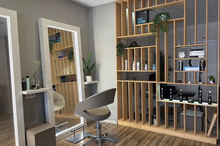 Un poste de coiffure avec un fauteuil gris face à un miroir encadré et éclairé par LED, une cloison-étagère à lattes de bois garnie de produits coiffants, une horloge et des plantes, sur un sol effet bois.